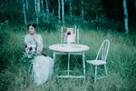 Bride sitting at white cake table in the woods